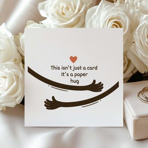 May include: A square white greeting card featuring a small red heart icon above the black text that reads, This isn't just a card it's a paper hug, accompanied by a graphic illustration of two dark brown arms reaching around to form a hug.