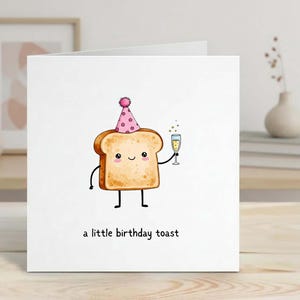Puede incluir: Una tarjeta de felicitación blanca con una ilustración de dibujos animados de una rebanada de pan tostado con un gorro de fiesta rosa y sosteniendo una copa de champán. El texto "a little birthday toast" está impreso debajo. La tarjeta está sobre una superficie de madera.