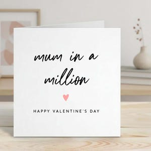 May include: A white greeting card with the text "mum in a million" in black script, a pink heart, and "HAPPY VALENTINE'S DAY" in black. The card is on a wooden surface.