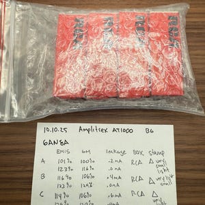 May include: A clear plastic bag containing a red, bubble-wrapped package. A handwritten paper with the text "10.10.25 Amplitrex AT1000 BG" and data tables is visible. The package is labeled with black text.