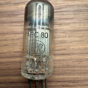 May include: A vintage vacuum tube with the text "FABC 80" and the "LORENZ" logo. The glass tube has a silver top and a clear body, revealing internal components. The tube has several pins at the base.