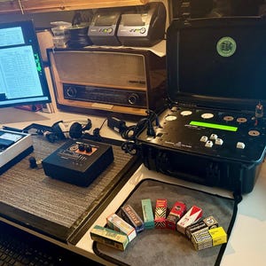 May include: A workspace with electronic equipment, including a black case with buttons and a green display, a vintage radio, and computer monitors displaying data. Several boxes of various brands are arranged in an arc.