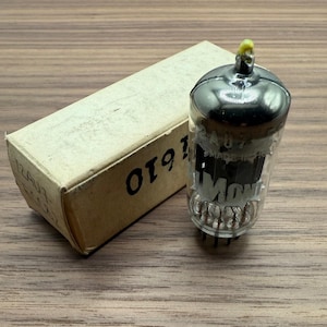 May include: A vintage vacuum tube with a clear glass body and a black top, next to a beige cardboard box. The box has the number "0191" printed on it. The tube has internal components visible through the glass.