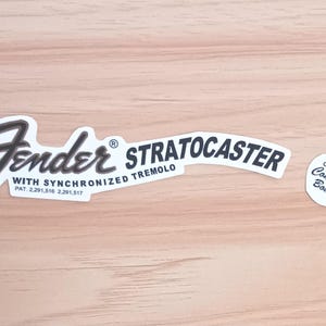 May include: Two white Fender Stratocaster guitar stickers with black text and a small white sticker with the text "Original Contour Body". The stickers are on a light brown wooden surface.