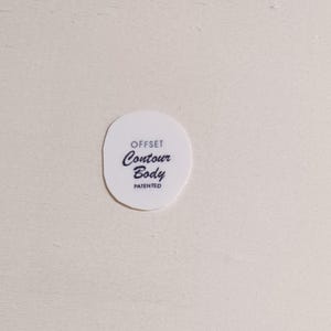 May include: A white, oval-shaped product with the words "OFFSET Contour Body PATENTED" printed in black. The text is arranged in three lines, with "Contour" in a cursive font. The product is set against a light-colored background.