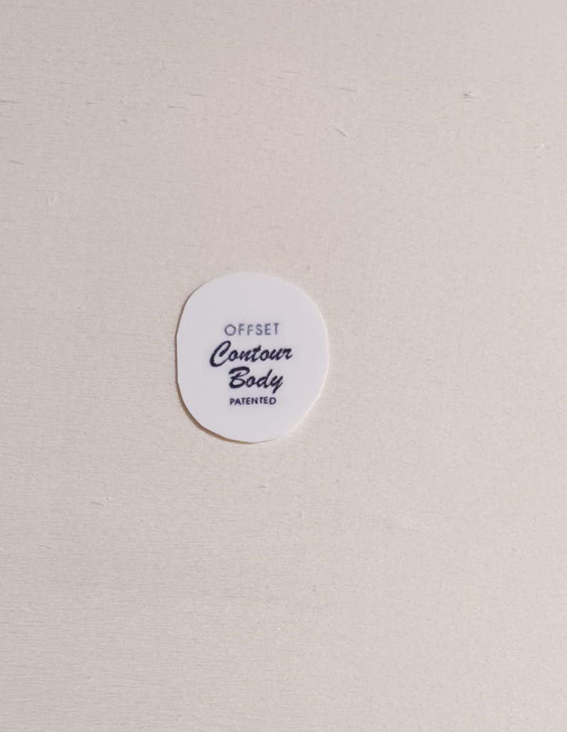 May include: A white, oval-shaped product with the words "OFFSET Contour Body PATENTED" printed in black. The text is arranged in three lines, with "Contour" in a cursive font. The product is set against a light-colored background.