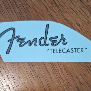 May include: A light blue decal featuring the Fender logo in a dark grey script font with the word TELECASTER printed in black capital letters underneath. The sticker has an irregular, cut-out shape with a smooth, matte finish on the surface.