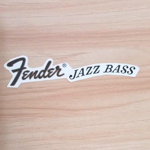 Może przedstawiać: Biała naklejka z czarnym logo Fender w charakterystycznym kroju pisma, po którym następuje tekst JAZZ BASS pogrubioną, czarną czcionką szeryfową. Naklejka zawiera mały symbol zastrzeżonego znaku towarowego obok nazwy Fender.