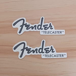 Könnte beinhalten: Zwei identische Aufkleber mit dem Fender-Markenlogo in einer dunkelgrauen Schreibschrift, platziert über dem Wort TELECASTER in einer einfachen schwarzen serifenlosen Schrift, alles auf weißem Hintergrund mit einem dünnen weißen Rand.