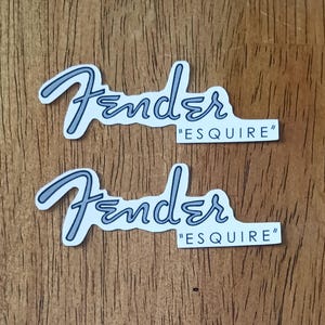 Może przedstawiać: Dwie naklejki Fender "ESQUIRE". Naklejki przedstawiają logo Fender w stylizowanej czcionce z jasnoniebieskim obrysem i ciemniejszym niebieskim wypełnieniem. Słowo "ESQUIRE" znajduje się w cudzysłowie pod logo.