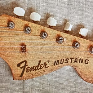 May include: Close-up of a Fender Mustang guitar headstock. The headstock is made of light brown wood, with the Fender logo and Mustang text in black. Six tuning pegs are visible, along with the strings.