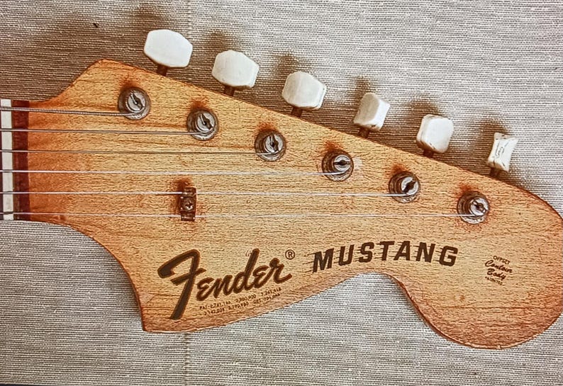 May include: Close-up of a Fender Mustang guitar headstock. The headstock is made of light brown wood, with the Fender logo and Mustang text in black. Six tuning pegs are visible, along with the strings.