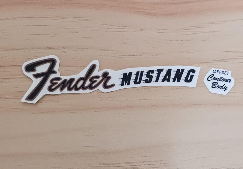 May include: A set of Fender Mustang guitar decals. The decals include the Fender logo, the word "Mustang", and a small decal that reads "Offset Contour Body". The decals are black and white with a brown outline.