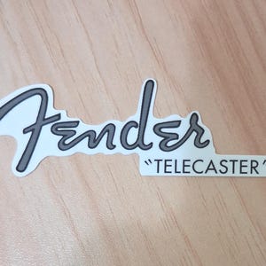 以下が含まれることがあります： 白い輪郭線で縁取られたグレーの筆記体フォントによるFenderブランドロゴと、その右側の白い長方形の枠内に黒い大文字でTELECASTERと印字されたデザインのダイカットステッカー。