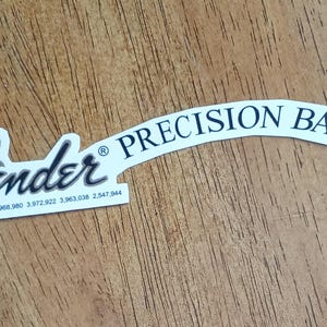 Może przedstawiać: Biała naklejka z czarno-szarym logo Fender i czarnym napisem "PRECISION BASS". Naklejka zawiera również numery patentów. Naklejka znajduje się na drewnianej powierzchni.