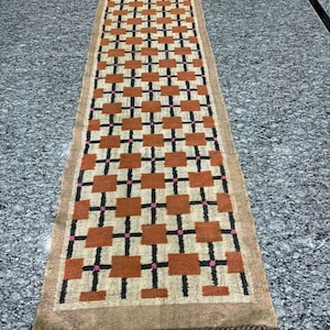 May include: A rectangular runner rug with a geometric pattern. The rug features a repeating pattern of rust-coloured squares, black lines, and small pink accents on a beige background. The rug has a brown border and fringe.