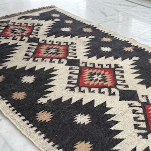 Pode incluir: Um tapete retangular tecido com um padrão geométrico repetitivo em preto, creme e vermelho. O design consiste em grandes formas de diamante com bordas serrilhadas, acentuadas por motivos menores em forma de estrela em bege e creme.