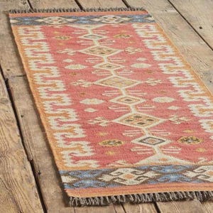 Handwoven wool jute kilim rug, 3x5 ft, coral-pink field with white geometric diamonds and blue-brown border, fringed ends.