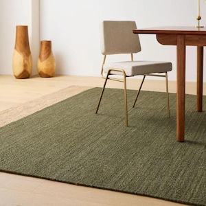 Puede incluir: Una alfombra rectangular verde con un borde beige, colocada sobre un suelo de madera clara. Una silla de comedor con asiento beige y patas doradas está sobre la alfombra. Dos jarrones de madera están en el fondo.