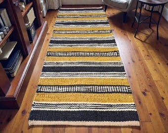 May include: A rectangular rug with horizontal stripes in black, white, and mustard yellow. The rug is placed on a wooden floor, and the design is visible from a high angle. The rug is a decorative element for a living space.