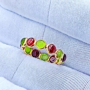 Könnte beinhalten: Ein Goldring mit einer Reihe von abwechselnd runden, in Zargenfassung gesetzten Cabochon-Edelsteinen in leuchtendem Rot und Limettengrün. Die Steine sind in einem durchgehenden Muster um den gesamten Umfang des polierten Goldrings angeordnet.