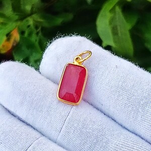 Könnte beinhalten: Ein rechteckiger Anhänger mit einem leuchtend rosa Edelstein, der in einen polierten goldfarbenen Metallrahmen gefasst ist. Der Anhänger verfügt über einen kleinen runden, goldfarbenen Verbindungsring an der Oberseite und ein klares Design.