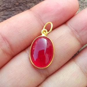 May include: A gold pendant featuring an oval, deep red gemstone. The gemstone is set in a gold bezel and has a polished, glossy finish. The pendant has a gold loop for a chain.