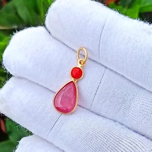 May include: A gold pendant featuring a small, round, bright red stone set above a larger, teardrop-shaped, faceted pink stone. The pendant includes a small circular gold jump ring at the top for attachment to a chain or necklace.
