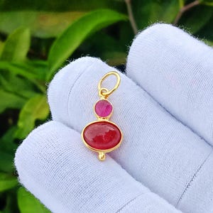 May include: A gold pendant featuring a small round pink gemstone positioned above a larger oval red gemstone. The pendant includes a gold jump ring at the top for attachment. The metal setting is yellow gold, providing a bright contrast to the stones.