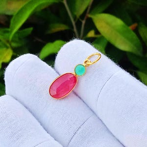 May include: A gold pendant featuring a small round mint green gemstone positioned above a larger oval-shaped vibrant pink gemstone. The pendant includes a small gold jump ring at the top for attachment, showcasing a polished metal setting.