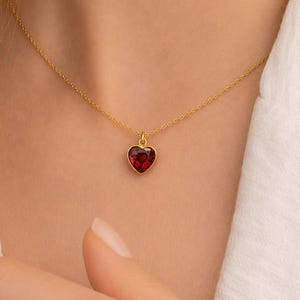Peut inclure: Un collier à fine chaîne dorée avec un petit pendentif en forme de cœur en pierre précieuse rouge à facettes. Le texte shreeshyamjewellerbp est visible dans le coin supérieur gauche. Le collier repose sur le cou avec un maillon simple.