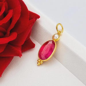 Puede incluir: Colgante de oro con un gran rubí ovalado y una pequeña perla. El rubí es de un color rojo-rosa vibrante, engastado en un bisel de oro. Tres cuentas doradas en la parte inferior. El colgante está sobre una superficie blanca.