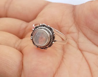 May include: A silver ring featuring a round, opalescent gemstone set within a decorative, dark-toned bezel. The ring has a thin, polished band. The gemstone has a milky appearance. The ring is shown on a hand.