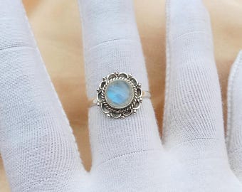 May include: A silver ring with a round, pale blue moonstone. The moonstone is set in an ornate, openwork bezel with a floral pattern. The ring is shown on a white surface.