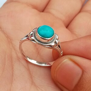 Può includere: Un anello d'argento con una pietra turchese rotonda e luminosa. L'anello presenta un design ornato attorno alla pietra e lungo la fascia. L'anello è mostrato su una mano.
