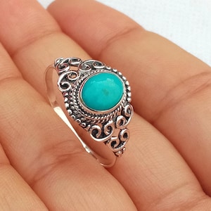Può includere: Un anello d'argento con una pietra ovale turchese brillante. La pietra è incastonata in una montatura decorativa in stile filigrana con intricati riccioli e dettagli perlati. L'anello è mostrato su uno sfondo chiaro.