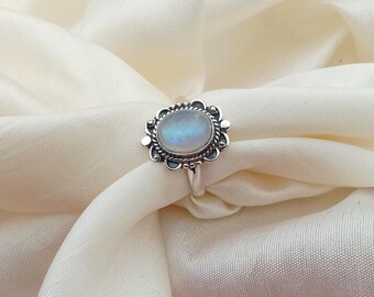 May include: A silver ring featuring an oval moonstone with an iridescent sheen, set within a decorative floral frame. The ring is presented on a cream-coloured fabric. The design highlights the ring's intricate details.