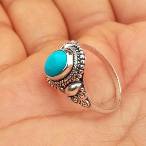 Può includere: Un anello d'argento con una pietra ovale turchese brillante. L'incastonatura dell'anello presenta dettagli intricati, tra cui un bordo perlato e accenti decorativi. L'anello è mostrato su uno sfondo chiaro.