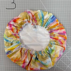 May include: A round, elasticized hair bonnet featuring a multicolored abstract brushstroke pattern in shades of yellow, pink, blue, and orange. The interior is lined with a solid white fabric and includes a small rectangular label with the text Just One Designs.