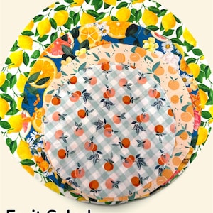 K&ouml;nnte beinhalten: Ein Set aus vier kreisf&ouml;rmigen Stoffsch&uuml;sseldeckeln, die in absteigender Gr&ouml;&szlig;e gestapelt sind. Die Muster zeigen gelbe Zitronen auf Wei&szlig;, Orangenscheiben auf Blau, Pfirsichillustrationen auf Pfirsichfarben und kleine Orangen auf gr&uuml;nem Vichy.