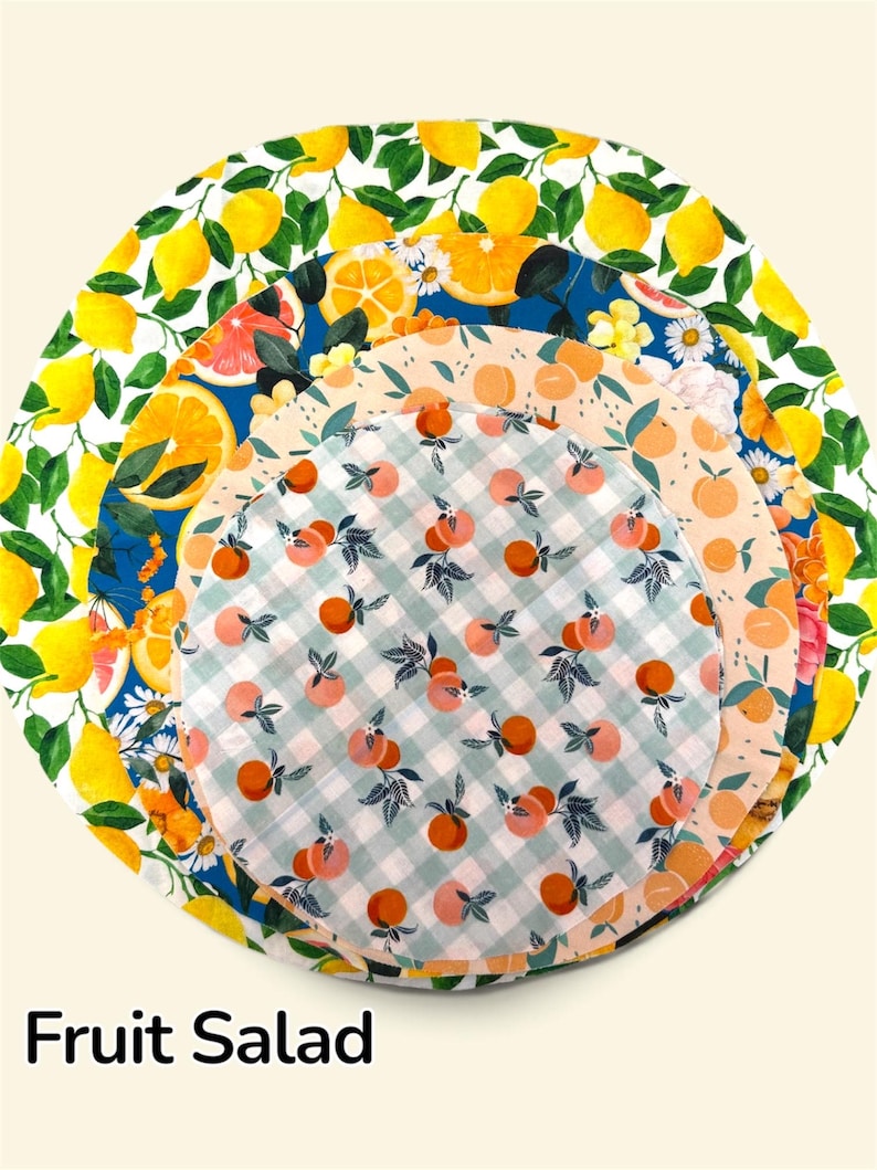 K&ouml;nnte beinhalten: Ein Set aus vier kreisf&ouml;rmigen Stoffsch&uuml;sseldeckeln, die in absteigender Gr&ouml;&szlig;e gestapelt sind. Die Muster zeigen gelbe Zitronen auf Wei&szlig;, Orangenscheiben auf Blau, Pfirsichillustrationen auf Pfirsichfarben und kleine Orangen auf gr&uuml;nem Vichy.