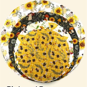 K&ouml;nnte beinhalten: Drei kreisf&ouml;rmige Stoffabdeckungen f&uuml;r Sch&uuml;sseln, die &uuml;bereinander liegen. Die gr&ouml;&szlig;te zeigt Sonnenblumen und V&ouml;gel auf Wei&szlig;. Die mittlere Schicht hat Pilze auf dunklem Grund. Die kleinste oberste Schicht zeigt Bienen und Tannenzapfen.