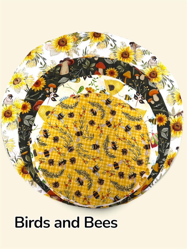 K&ouml;nnte beinhalten: Drei kreisf&ouml;rmige Stoffabdeckungen f&uuml;r Sch&uuml;sseln, die &uuml;bereinander liegen. Die gr&ouml;&szlig;te zeigt Sonnenblumen und V&ouml;gel auf Wei&szlig;. Die mittlere Schicht hat Pilze auf dunklem Grund. Die kleinste oberste Schicht zeigt Bienen und Tannenzapfen.