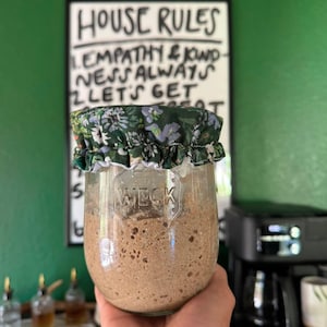 May include: A clear glass Weck jar filled with a bubbly, light brown sourdough starter. The jar is topped with a fabric cover featuring a dark green background with a small, repeating floral pattern in shades of blue, white, and light green.