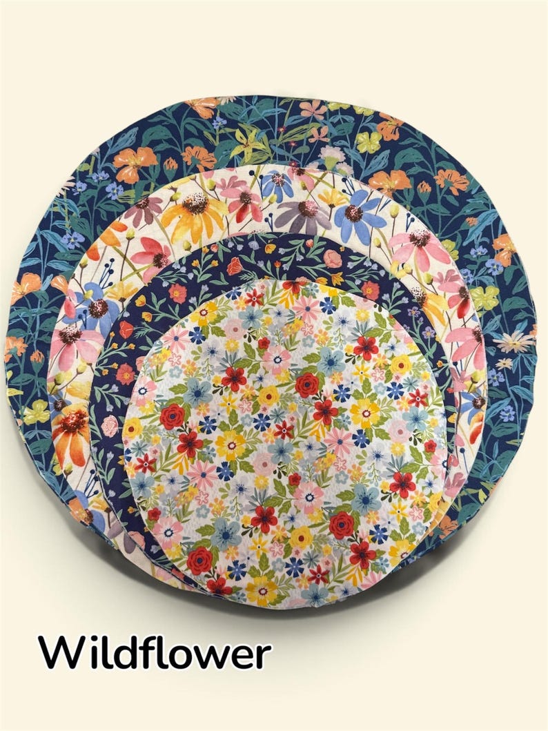 K&ouml;nnte beinhalten: Ein Set aus vier ineinander verschachtelten, kreisf&ouml;rmigen Sch&uuml;sseldeckeln aus Stoff mit verschiedenen Blumenmustern in den Farben Marineblau, Orange, Gelb, Rosa und Rot. Der Text "Wildflower" ist unten im Bild in Schwarz aufgedruckt.