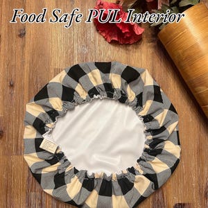 K&ouml;nnte beinhalten: Eine runde Sch&uuml;sselabdeckung mit einem schwarz-beigen Karomuster auf der Au&szlig;enseite und einer glatten, lebensmittelechten wei&szlig;en PUL-Innenseite. Der Text 100% cotton exterior und Food Safe PUL Interior ist &uuml;ber der runden Stoffabdeckung zu sehen.