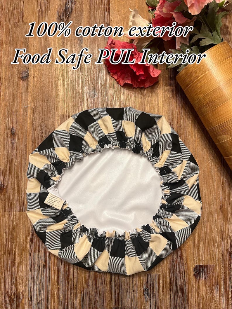 K&ouml;nnte beinhalten: Eine runde Sch&uuml;sselabdeckung mit einem schwarz-beigen Karomuster auf der Au&szlig;enseite und einer glatten, lebensmittelechten wei&szlig;en PUL-Innenseite. Der Text 100% cotton exterior und Food Safe PUL Interior ist &uuml;ber der runden Stoffabdeckung zu sehen.