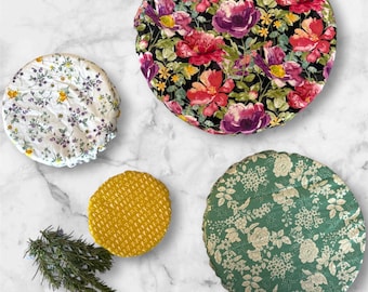 May include: Four round fabric bowl covers with floral patterns. One cover has a black background with red and purple flowers. Another has a white background with purple and yellow flowers. A third is green with white flowers, and the last is yellow.