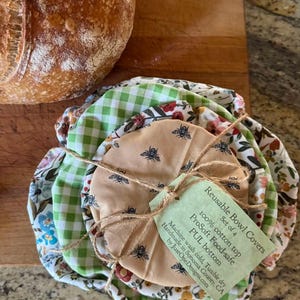 K&ouml;nnte beinhalten: Ein Set aus vier wiederverwendbaren Sch&uuml;sselabdeckungen mit verschiedenen Stoffmustern, darunter gr&uuml;nes Vichy-Karo, Blumenmuster und ein beiger Stoff mit Bienenmotiv. Ein hellgr&uuml;nes Etikett mit Bindfaden tr&auml;gt die Aufschrift: Reusable Bowl Covers, Set of 4, 100% cotton top, ProSoft Food-safe, PUL bottom.