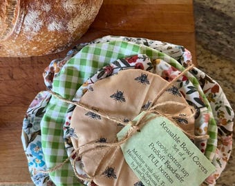 Op de afbeelding: Een set van vier herbruikbare komafdekkingen met diverse stofpatronen, waaronder groen gingham, bloemenprints en een beige stof met een bijenmotief. Een lichtgroen label vastgebonden met touw vermeldt: Reusable Bowl Covers, Set of 4, 100% cotton top, ProSoft Food-safe, PUL bottom.